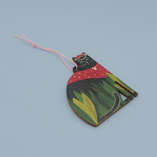 A wooden decoration shaped like a cat with black fur, green leaves, a red cape, and flowers on it's head. The decoration is hanging on a pink string and is on a blue background.