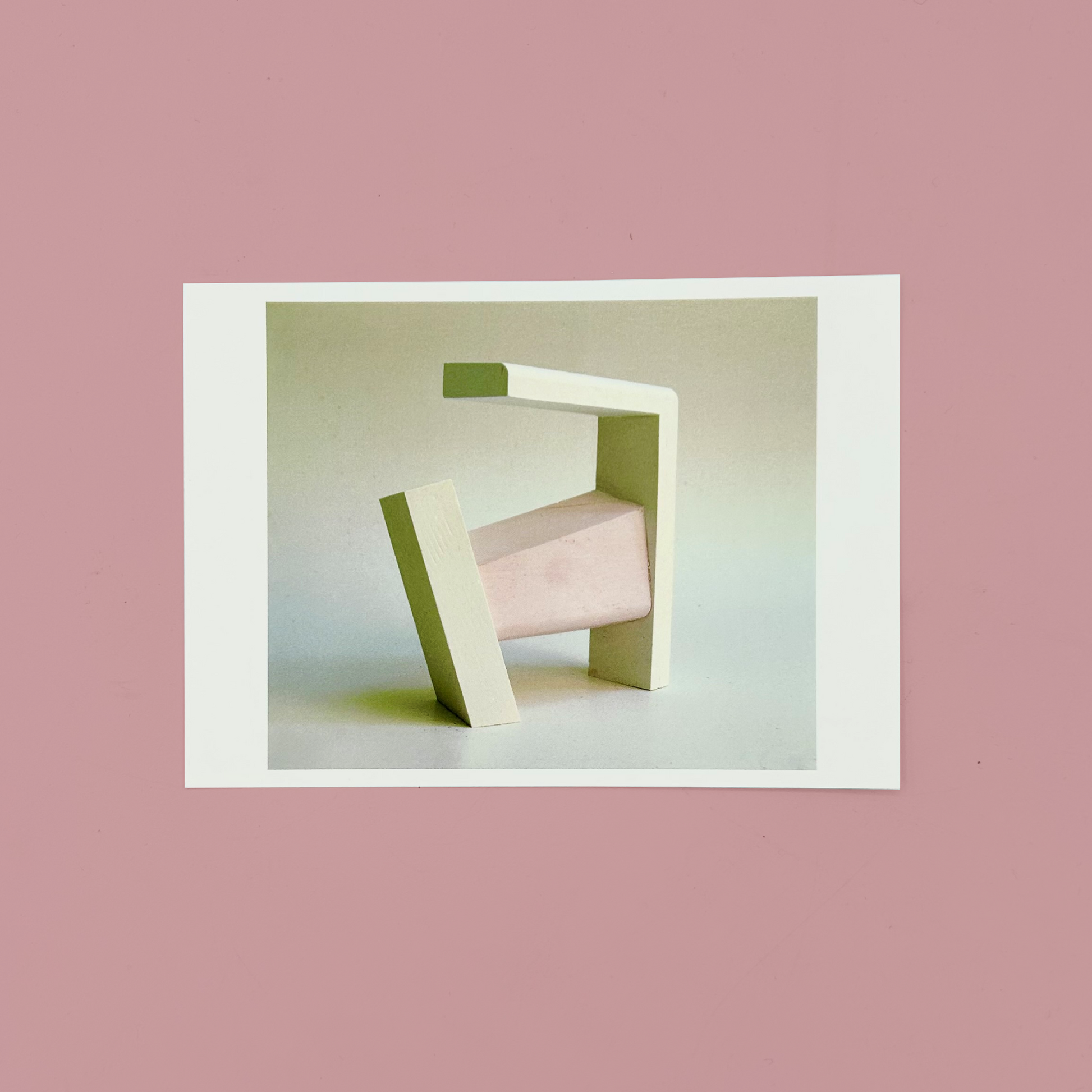 Modern sculpture of a geometric design on a white postcard. The postcard is on a pink background.