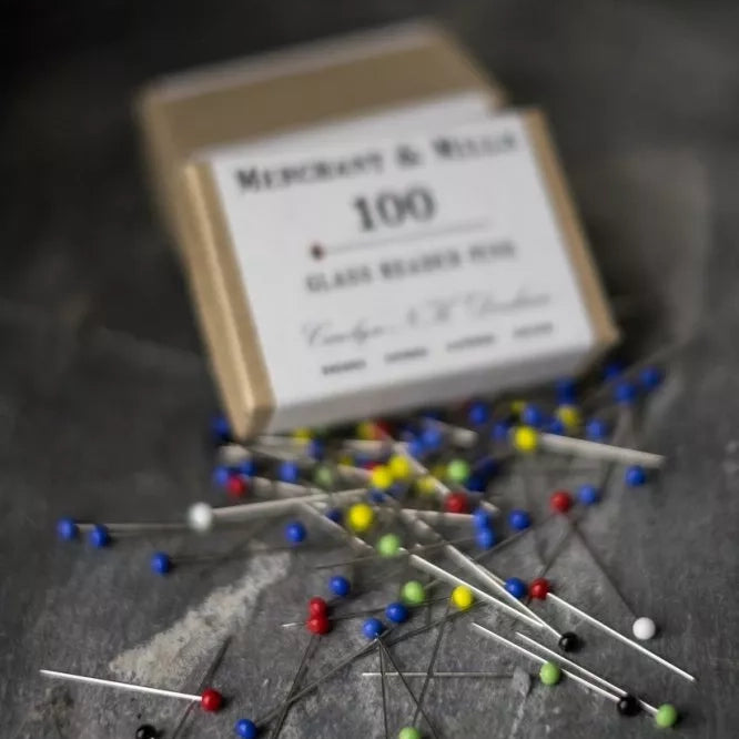 Box of multicolour glass headed pins with scattered pins on a dark surface