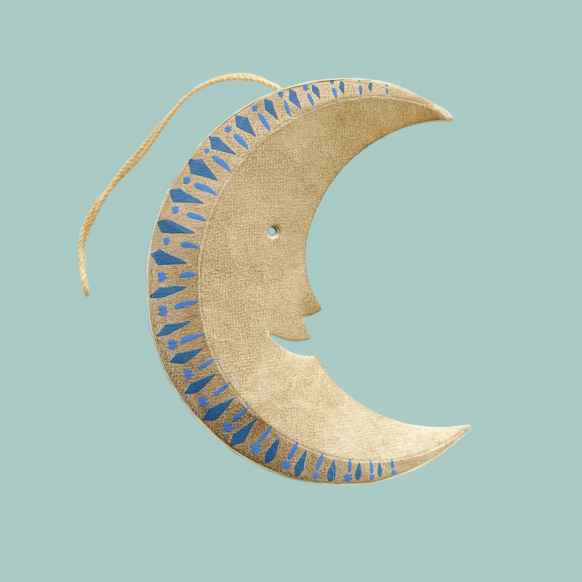 A crescent moon hanging decoration made from recycled paper, featuring a stylized face profile and blue patterned accents, suspended by a gold string against a solid light blue background.
