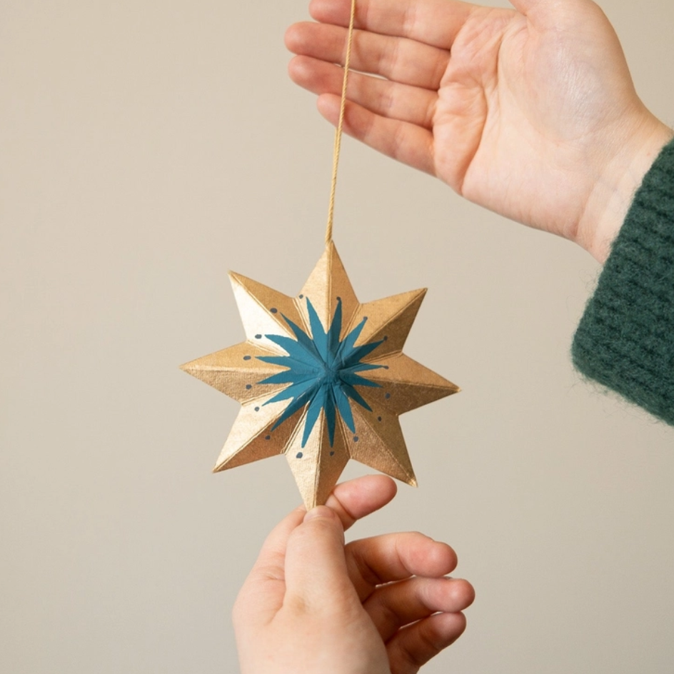 A handcrafted wooden eight-pointed star ornament with a blue starburst center and gold glitter accents, held by two hands against a neutral beige background.