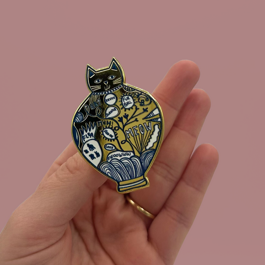 A hand holding an enamel pin of a cat shaped vase.