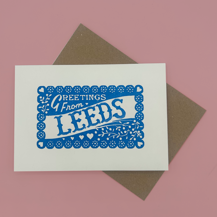 A white greeting card with a blue decorative border and the words 'Greetings From LEEDS' in blue, placed on top of a brown envelope against a pink background.