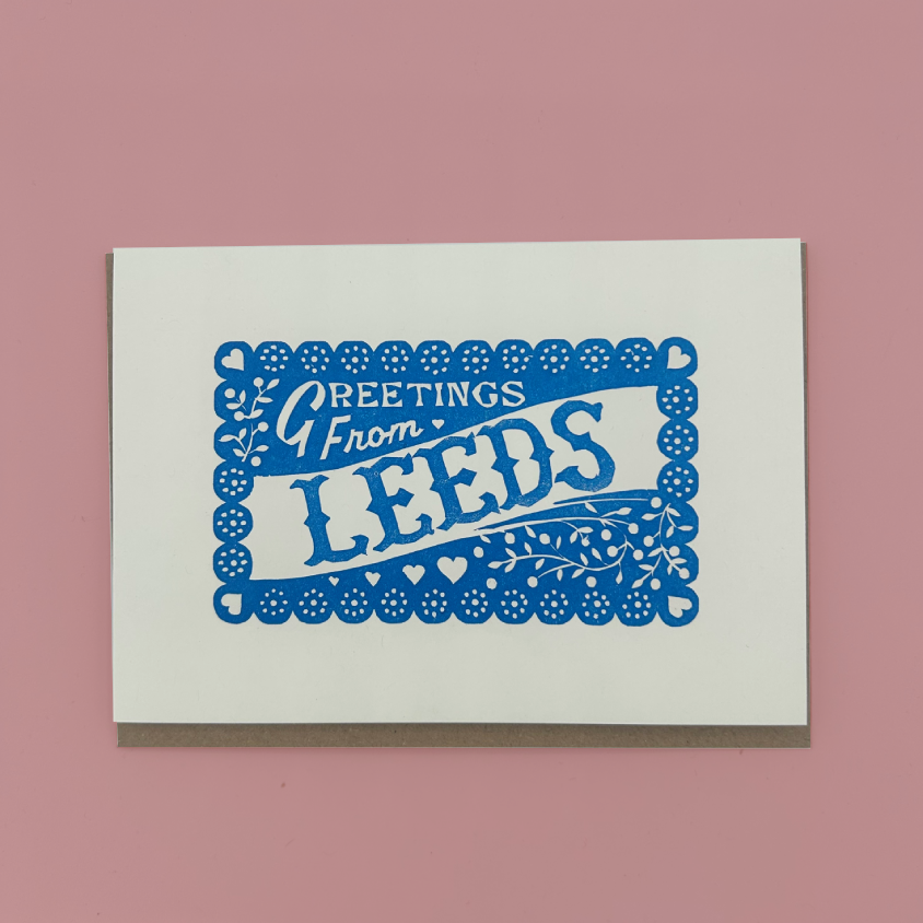 A square white greeting card with a blue decorative border and the words 'Greetings From LEEDS' in blue, photographed against a solid pink background.