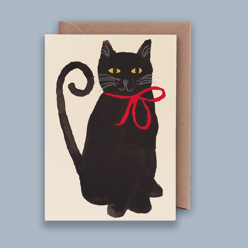 A greeting card featuring a painted black cat with yellow eyes and a red bow around its neck, shown with a brown kraft envelope against a muted blue-gray background.