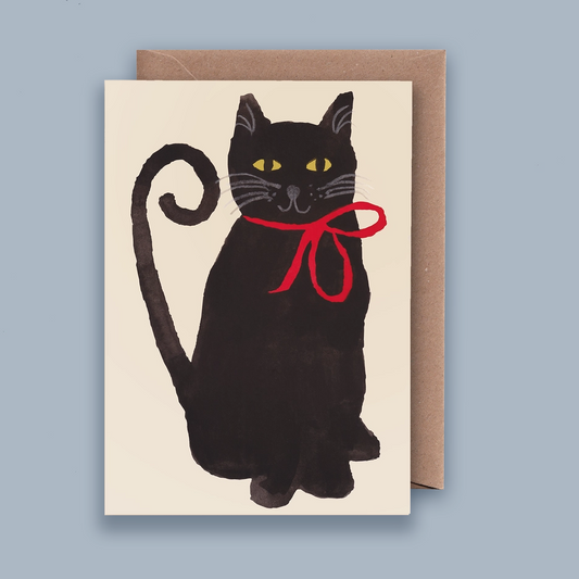 A greeting card featuring a painted black cat with yellow eyes and a red bow around its neck, shown with a brown kraft envelope against a muted blue-gray background.
