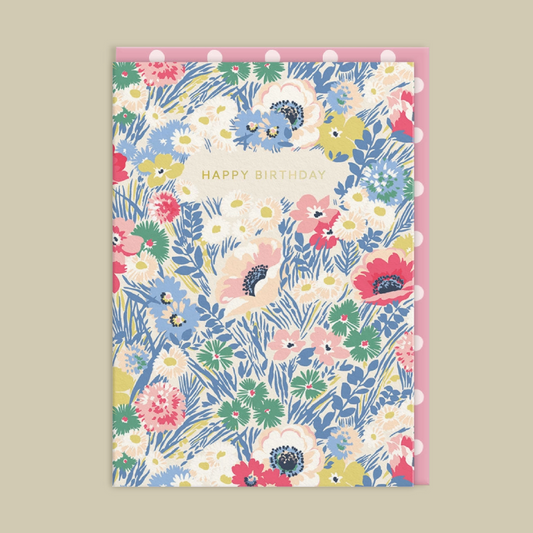 A floral birthday card featuring a multicolor pattern of flowers and green leaves on a cream background with the words 'HAPPY BIRTHDAY' in gold. It is shown with a matching pink envelope on a solid beige background.