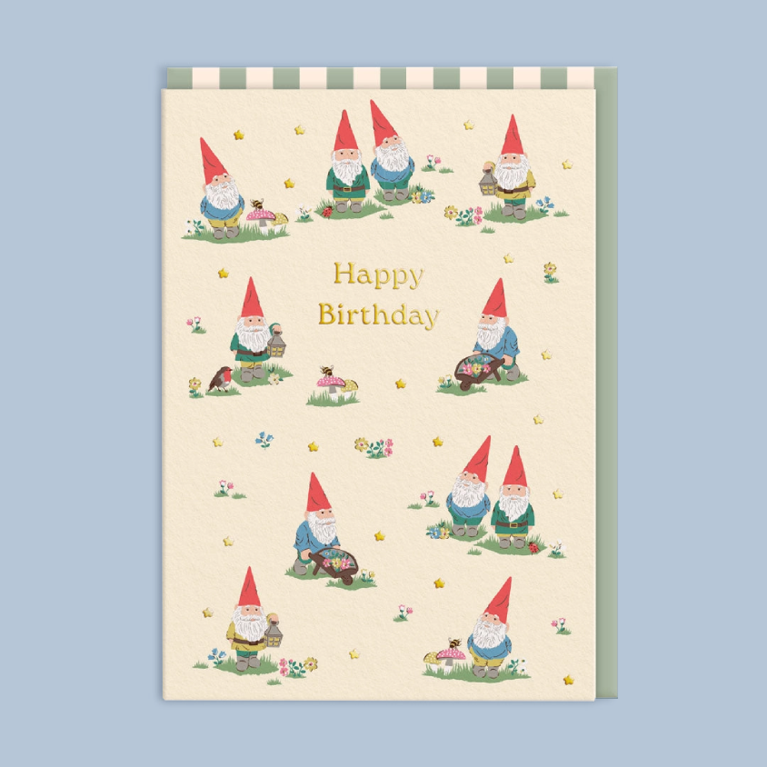 A rectangular birthday card with a cream background, decorated with multiple cute garden gnome illustrations and the words 'Happy Birthday' in gold lettering. The card is shown against a solid light blue background.