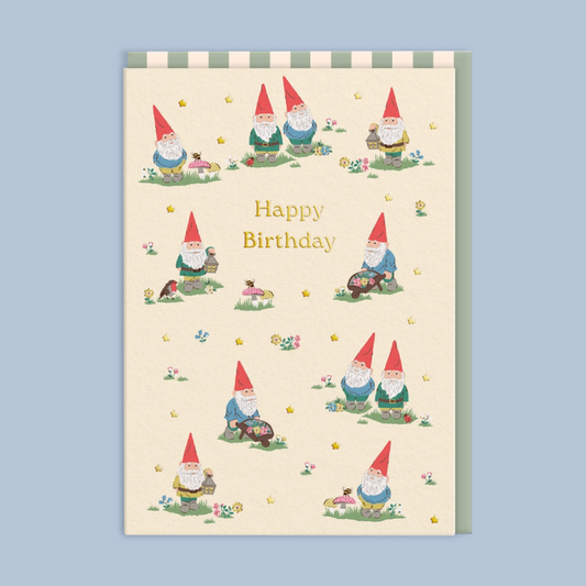 A rectangular birthday card with a cream background, decorated with multiple cute garden gnome illustrations and the words 'Happy Birthday' in gold lettering. The card is shown against a solid light blue background.