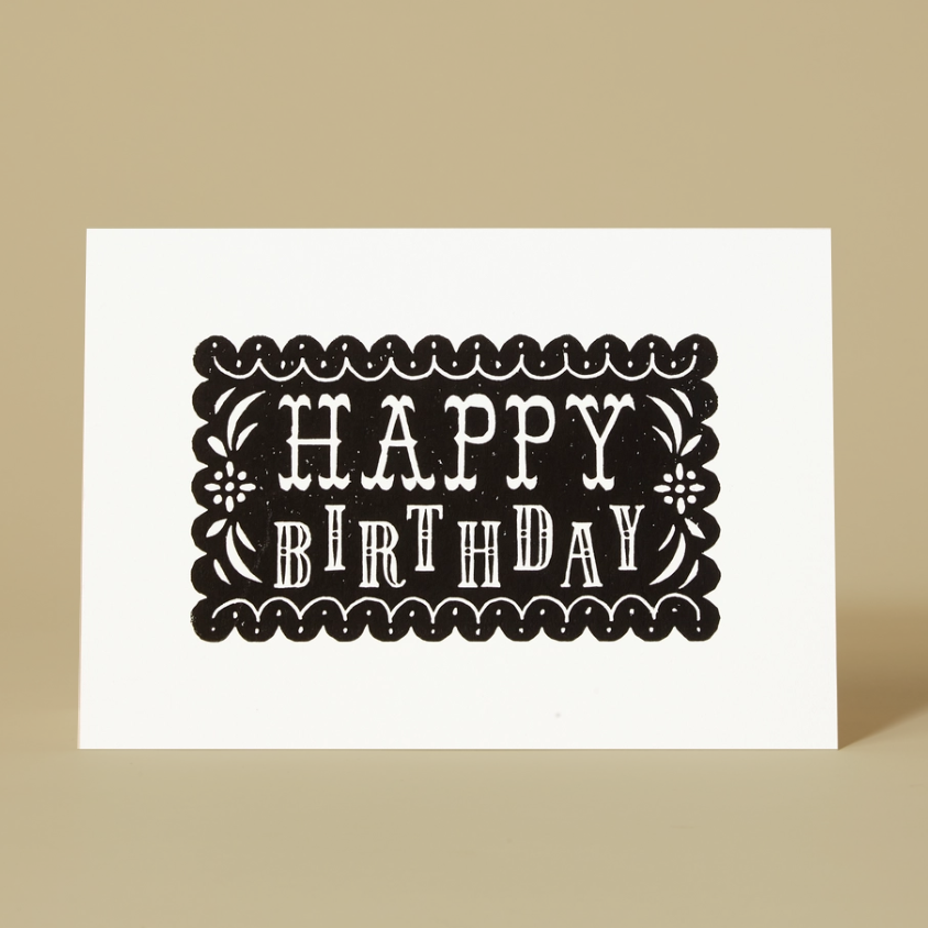 A white landscape orientation greeting card standing upright on a beige background, printed with the words “HAPPY BIRTHDAY” in white inside a black decorative border.