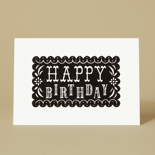 A white landscape orientation greeting card standing upright on a beige background, printed with the words “HAPPY BIRTHDAY” in white inside a black decorative border.