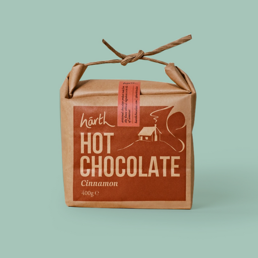A square kraft paper package of Harth Hot Chocolate Cinnamon, featuring a brown label with white text and graphics, tied with a twine handle on a solid light green background.
