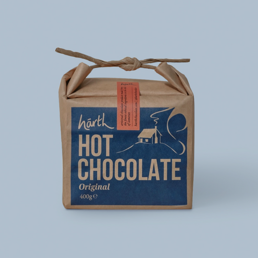 A kraft paper gift box tied with a twine handle, featuring a blue front label that reads 'harth HOT CHOCOLATE Original 400g' with a cream illustration of a house and cocoa bean. A small orange tag is attached on the side, set against a solid light blue background.