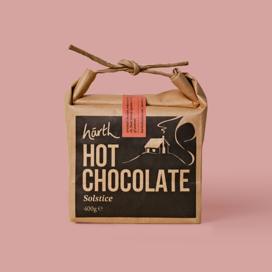 A square kraft paper gift box tied with a twine handle, featuring a black front label that reads 'narth HOT CHOCOLATE Solstice 400g e'. The box is photographed on a solid dusty pink background.