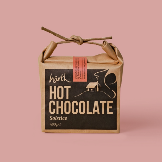A square kraft paper gift box tied with a twine handle, featuring a black front label that reads 'narth HOT CHOCOLATE Solstice 400g e'. The box is photographed on a solid dusty pink background.