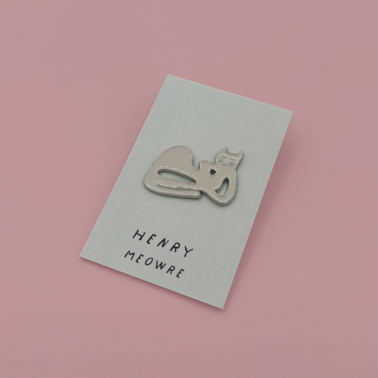 A silver metal enamel pin shaped like a sleeping cat, mounted on a white display card with the text 'HENRY MEOWRE', set against a solid pink background.