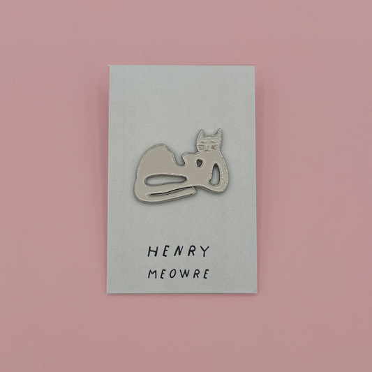 A silver metal enamel pin shaped like a sleeping cat, mounted on a light blue-gray backing card printed with the text 'HENRY MEOWRE'. The card is centered on a solid pink background.