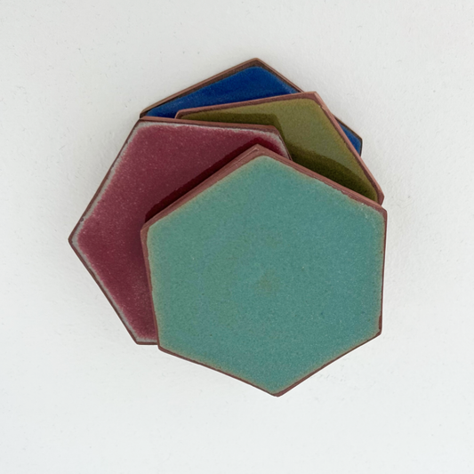 Four hexagonal ceramic coasters stacked in a fan arrangement on a plain white background. The front piece is a muted teal-green, with a dusty rose piece behind it, and blue and olive green pieces layered further back.