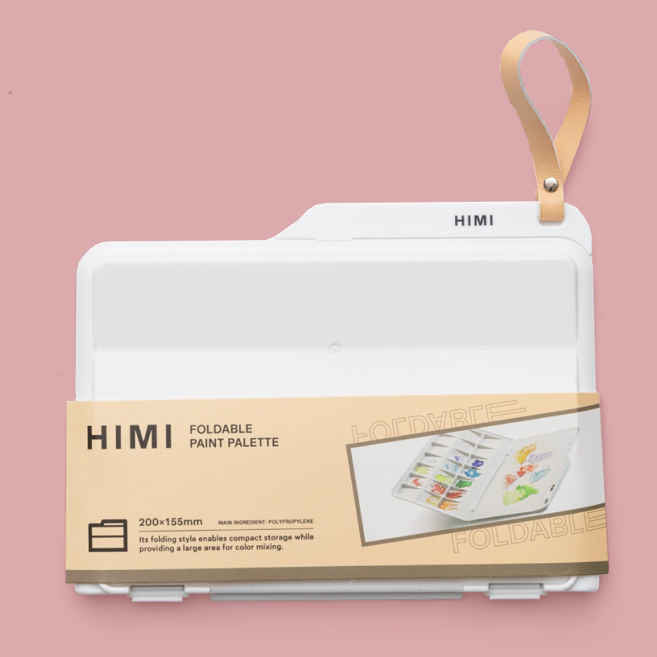 A HIMI foldable paint palette in white, shown folded with a tan carrying strap and a tan paper band around the lower half. The background is a solid dusty pink.