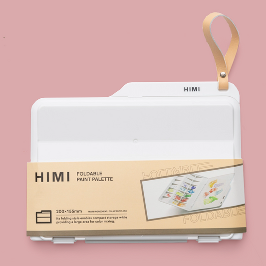 A HIMI foldable paint palette in white, shown folded with a tan carrying strap and a tan paper band around the lower half. The background is a solid dusty pink.