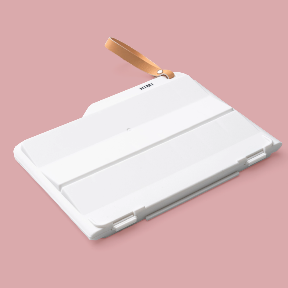 A white paint palette with a tan wrist strap, shown at an angle on a solid pink background. The brand name 'HIM' is printed in black on the top surface.