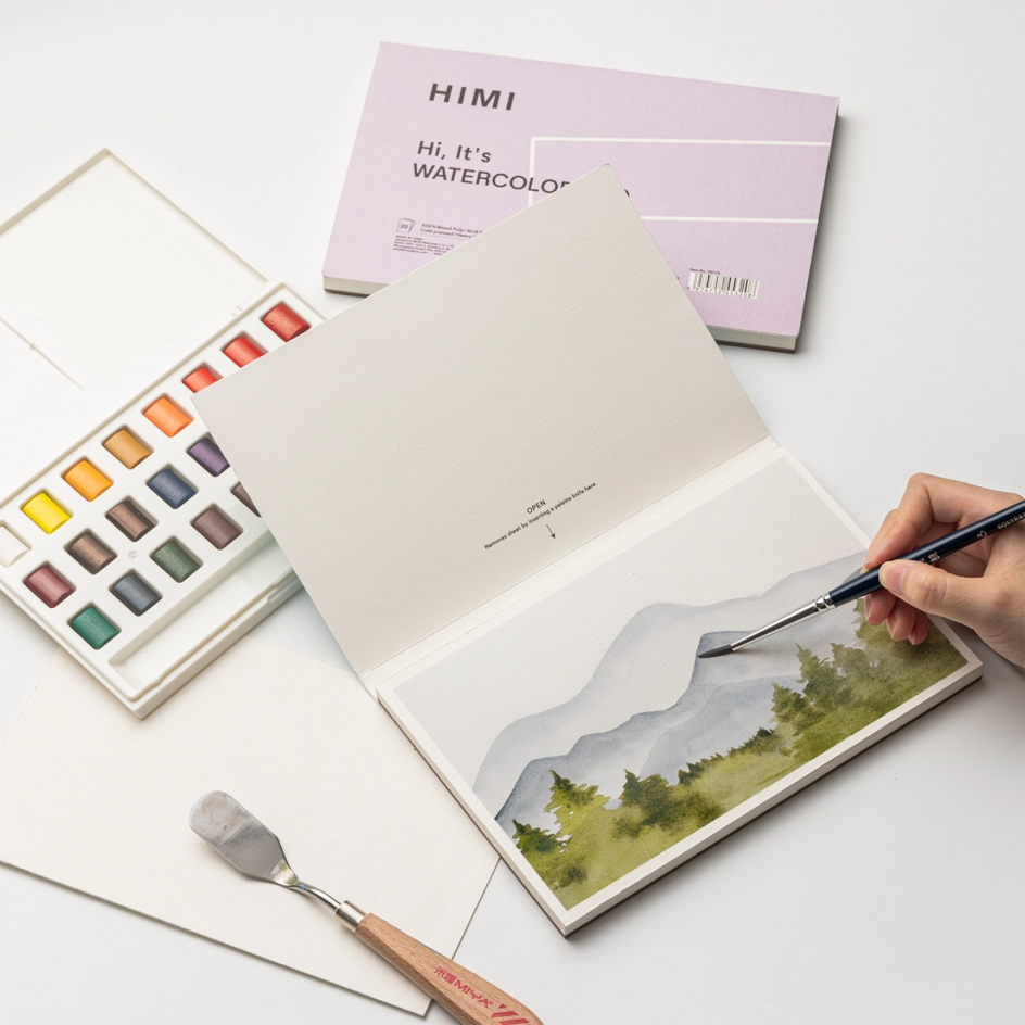 A watercolour painting set on a white surface, featuring an open watercolor pad with a hand painting a landscape, a watercolour palette with multiple pans, a paper pad labeled HIMI, and a paint palette knife.