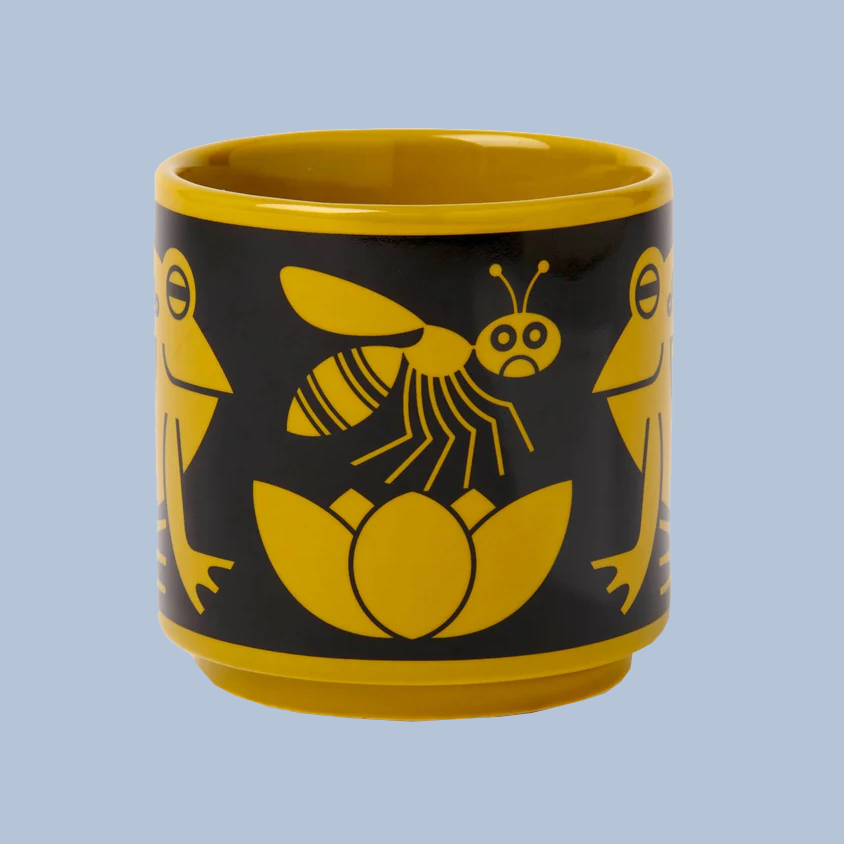 A yellow and black ceramic cup featuring a whimsical design of an insect, lotus flower, and stylized faces on the sides, set against a solid light blue background.