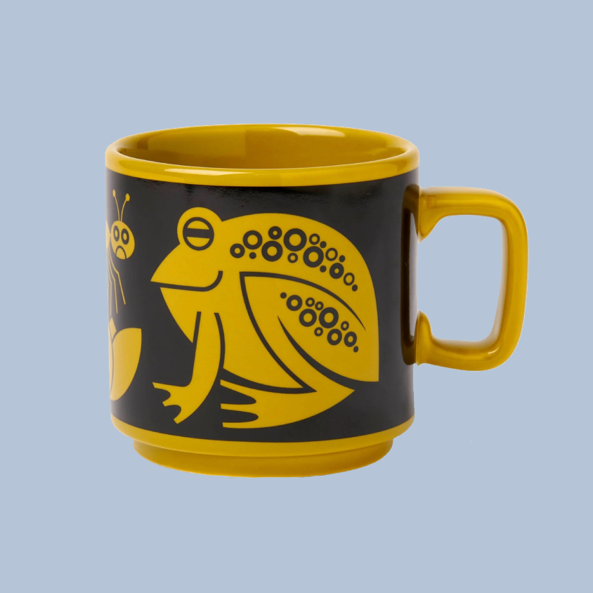 A yellow and black ceramic mug featuring a stylized yellow frog and insect illustration on a light blue background.