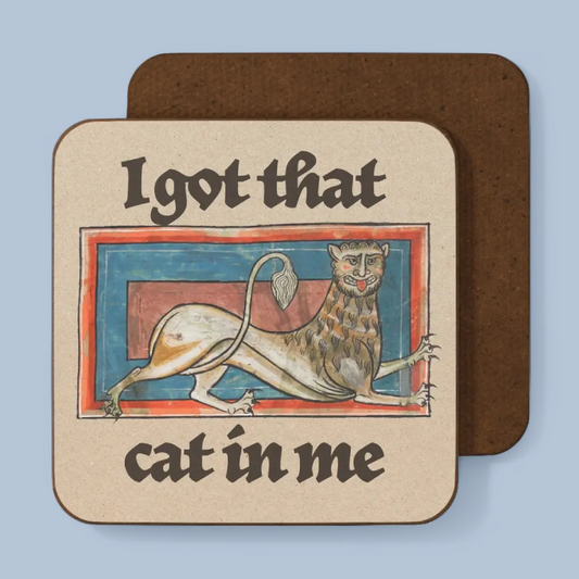 A square coaster with rounded corners shown on a light blue background. The front of the coaster has a beige printed design with the text 'I got that cat in me' and an illustration of a stylized cat in a medieval-style frame.