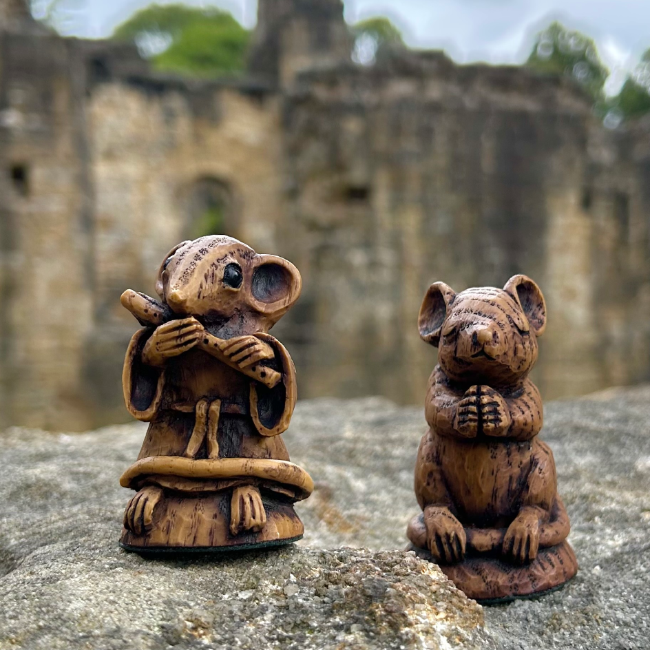 Two small carved mouse figurines with a natural wood grain finish, one holding a flute and the other with its paws together, displayed on a stone surface with a blurred Kirkstall Abbey in the background.