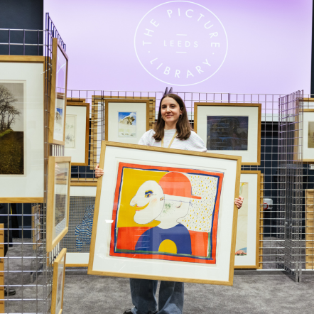 Person holding a framed artwork with a colorful design, surrounded by other framed pictures in a store setting.