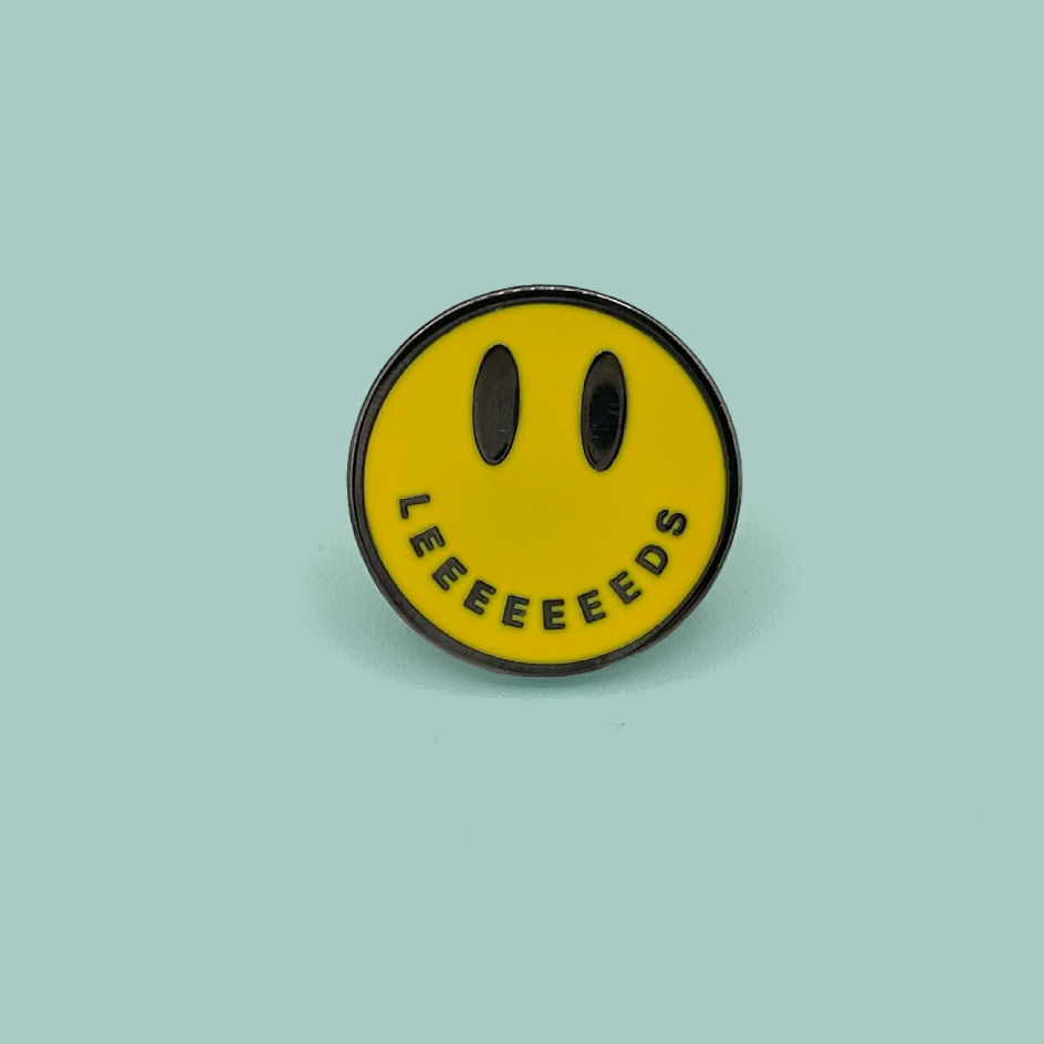 A circular yellow enamel pin featuring a minimalist black smiley face with the word 'LEEEEEEEDS' curved along the bottom edge, set against a solid light teal background.