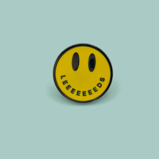 A circular yellow enamel pin featuring a minimalist black smiley face with the word 'LEEEEEEEDS' curved along the bottom edge, set against a solid light teal background.