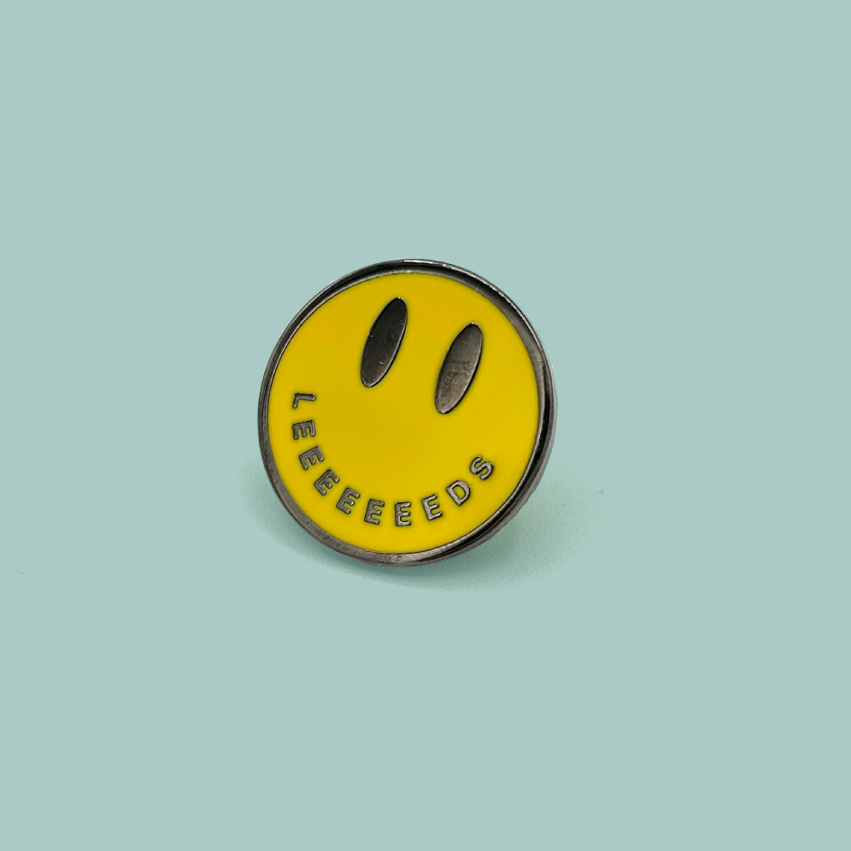A circular yellow enamel pin featuring a minimalist black smiley face with the word 'LEEEEEEEDS' curved along the bottom edge, set against a solid light teal background.