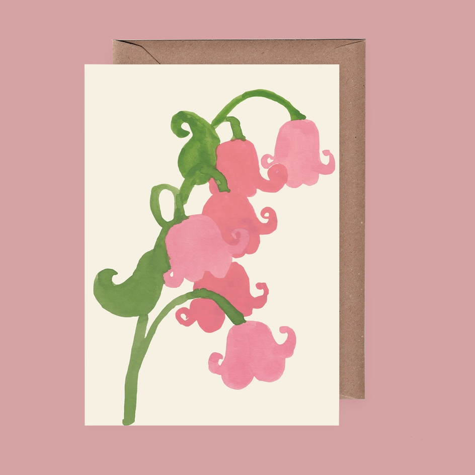 A vertical greeting card with a watercolor illustration of pink lily-of-the-valley flowers on a cream background, shown with a brown kraft envelope behind it on a dusty pink backdrop.