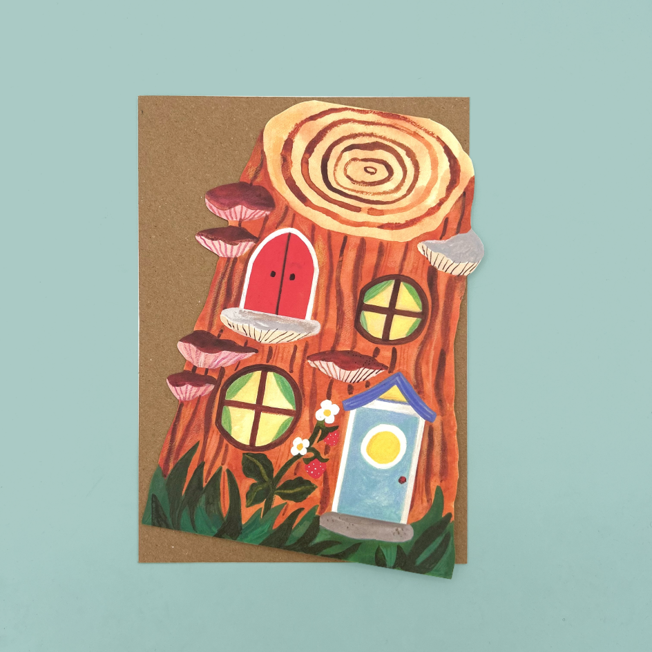 A fairy house card featuring a whimsical illustration of a tree stump with mushrooms, a small blue house, and a red door, displayed on a light blue background.