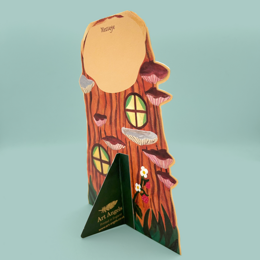 A decorative greetings card shaped like a tree trunk with mushroom shelves, circular window cutouts, small flowers, and a triangular base printed with 'Art Angels' and 'The Art of Living'.