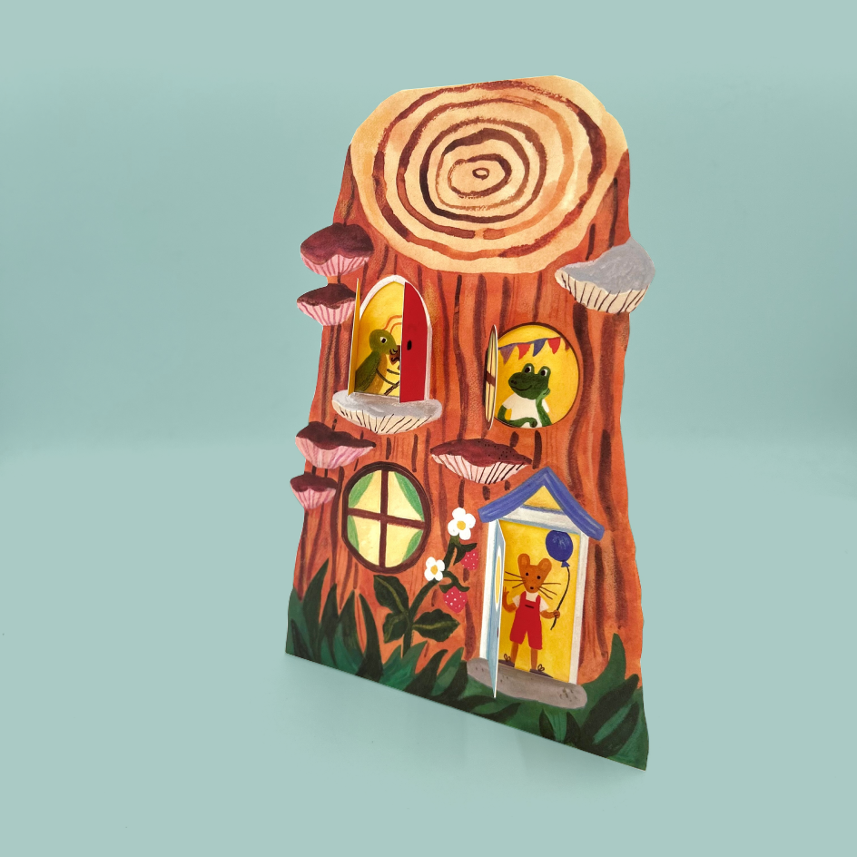 A whimsical greetings card shaped to look like a tree trunk, featuring colorful frog characters in circular windows, mushroom details, and a small blue-trimmed door, set against a solid light teal background.
