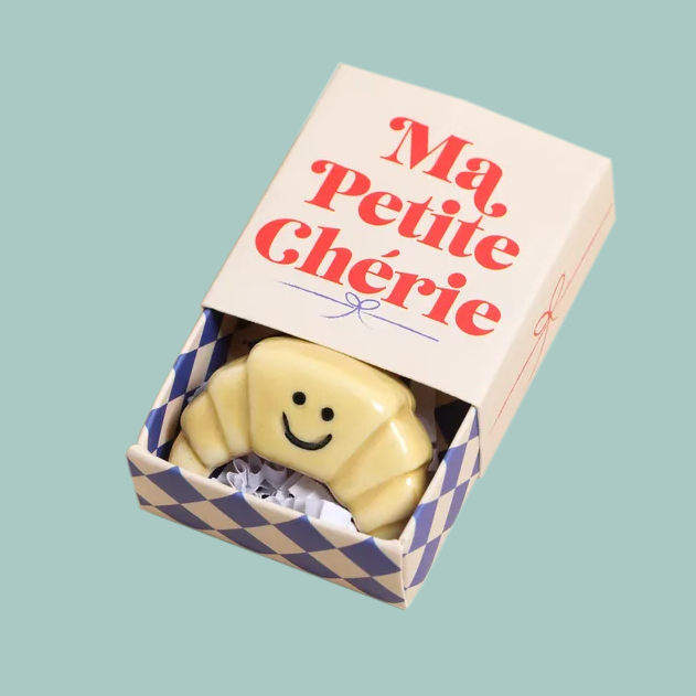 A small ceramic croissant token with a smiley face, inside a matchbox with a checked inside and 'ma petite cherie' written on the lid.