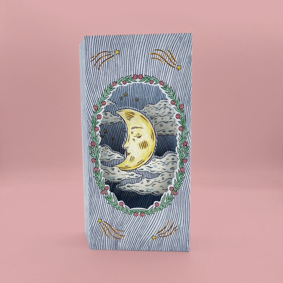 A tall greetings card with a whimsical celestial illustration, standing upright against a solid pink background. The package features a blue and white striped border with a central oval illustration of a smiling crescent moon, stars, and clouds, framed by a green wreath with red berries.