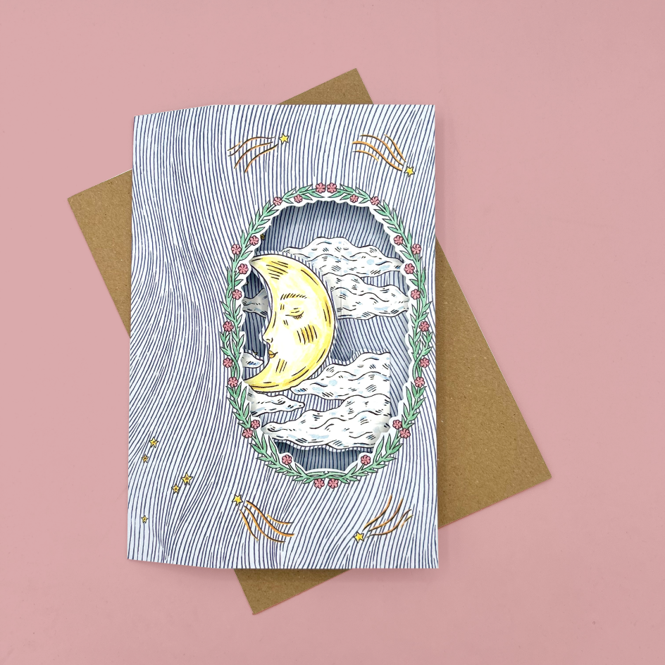 An illustrated card with a crescent moon and clouds, placed on top of a brown envelope against a pink background.