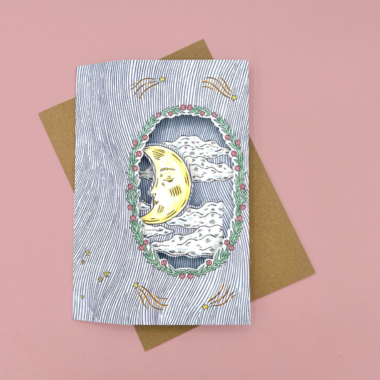 An illustrated card with a crescent moon and clouds, placed on top of a brown envelope against a pink background.