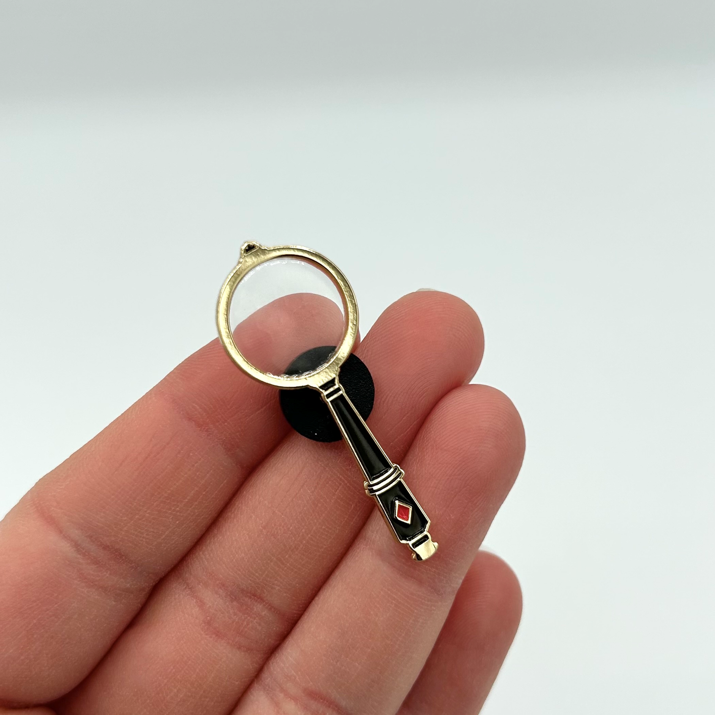Magnifying glass pin badge held on a hand against a light background.