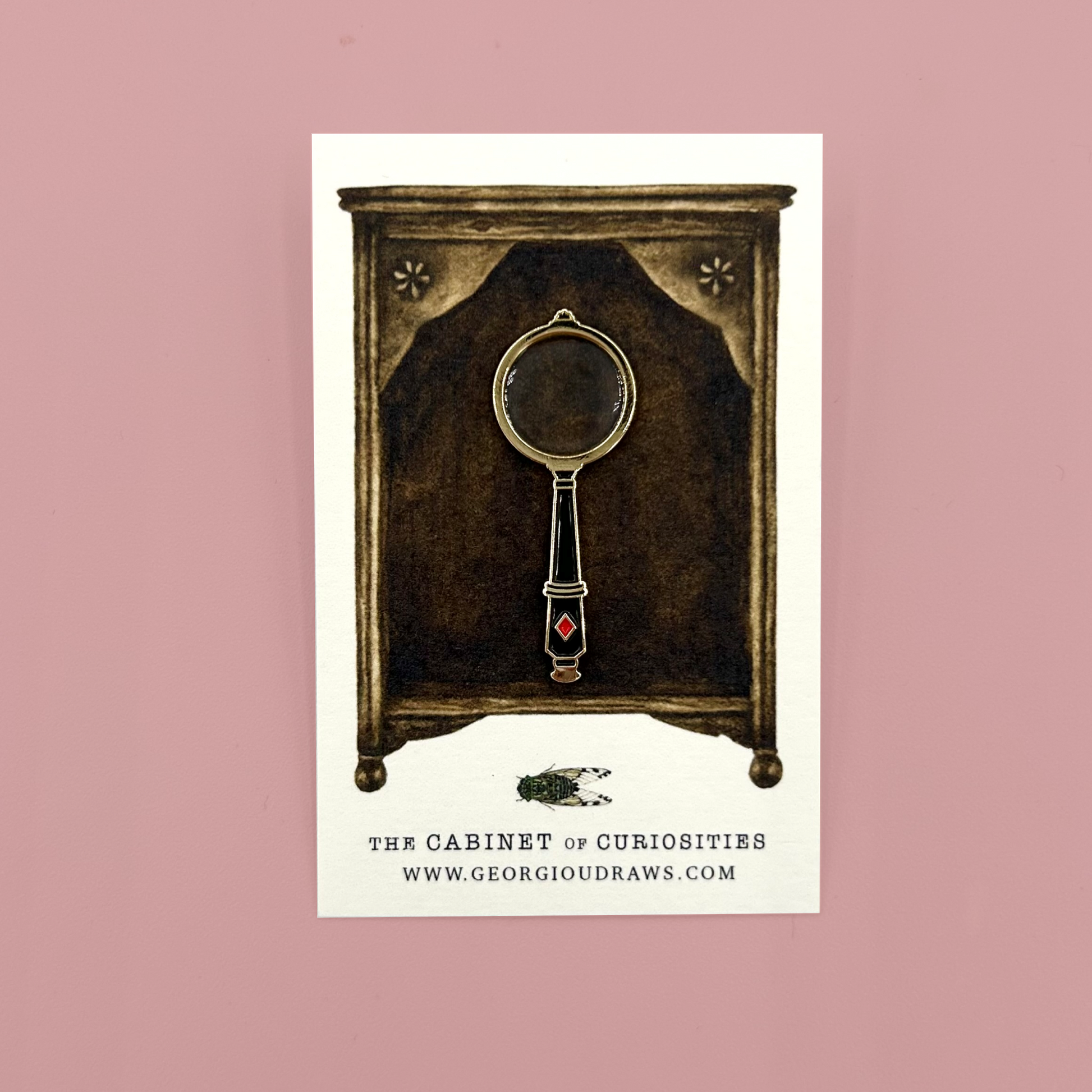 Illustrated backing card of a cabinet of curiosities with a vintage-style magnifying glass pin badge on a pink background.
