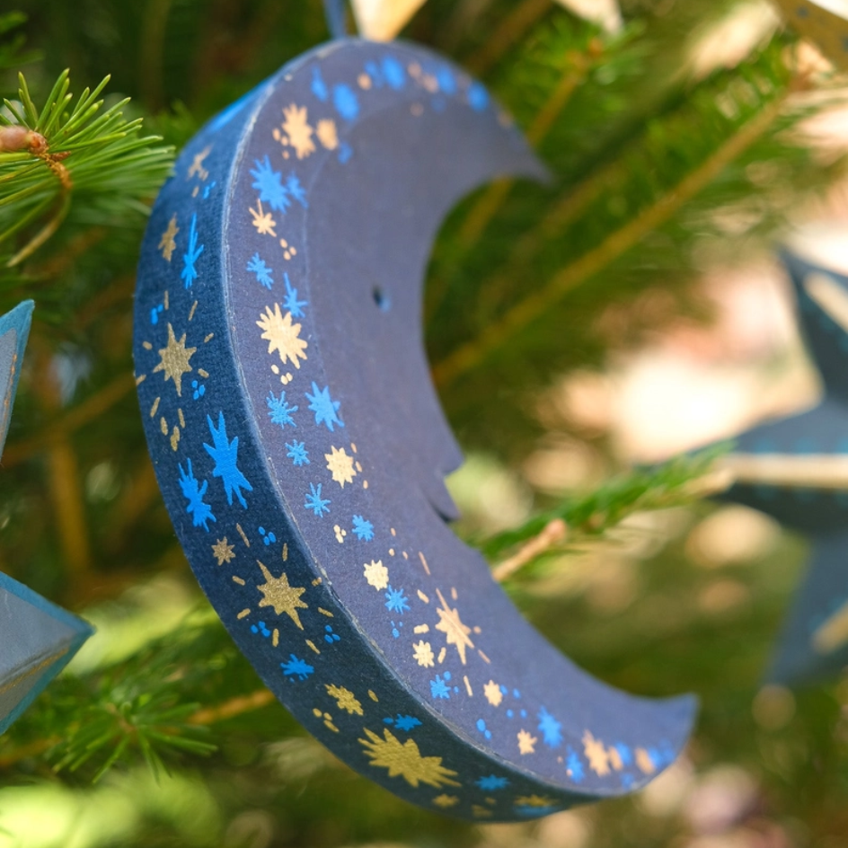 A crescent moon Christmas ornament painted in dark blue with gold and light blue star patterns, hanging on a green pine tree branch.