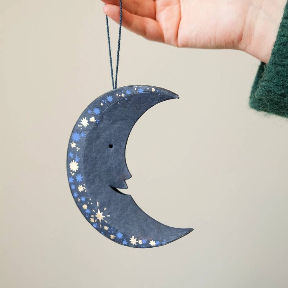 A hand holds a dark blue crescent moon hanging ornament decorated with gold and light blue stars and snowflakes against a plain beige background.