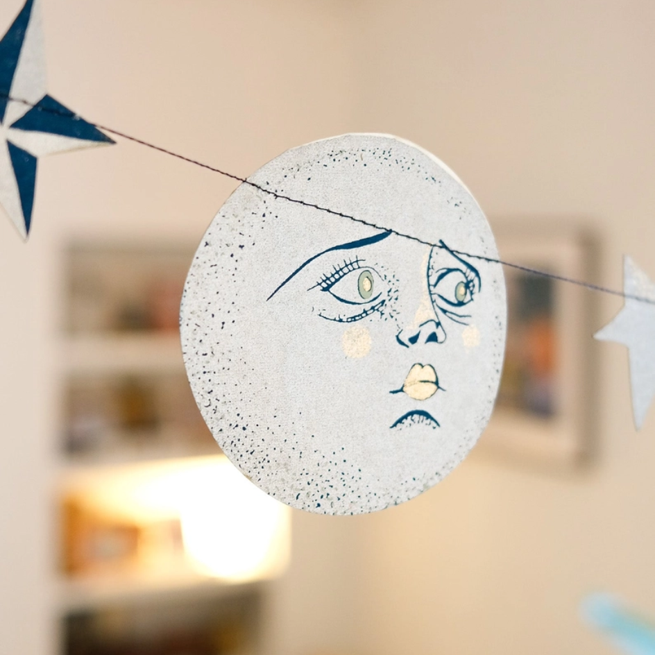 A circular moon-shaped garland decoration with a speckled light gray surface and a stylized face in dark blue and cream, suspended by a string in a softly blurred indoor setting.