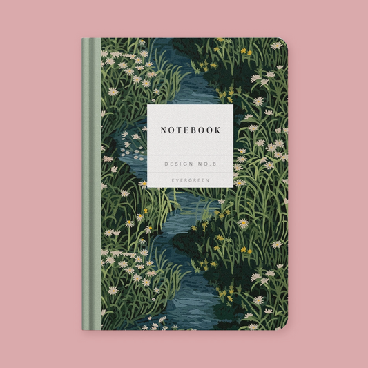 A notebook with a nature illustration cover featuring a winding blue stream surrounded by green grasses and white wildflowers, with a white label on the front reading 'NOTEBOOK', 'DESIGN NO.8', and 'EVERGREEN'.