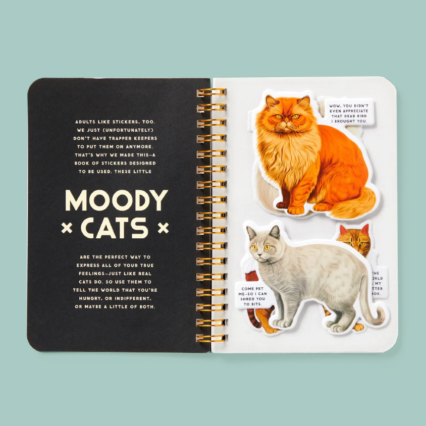 An open spiral-bound sticker book titled 'MOODY CATS' on a solid light blue background. The left page is black with white text, and the right page features die-cut cat stickers including an orange tabby, a white cat, and a small orange tabby with a red bow tie.