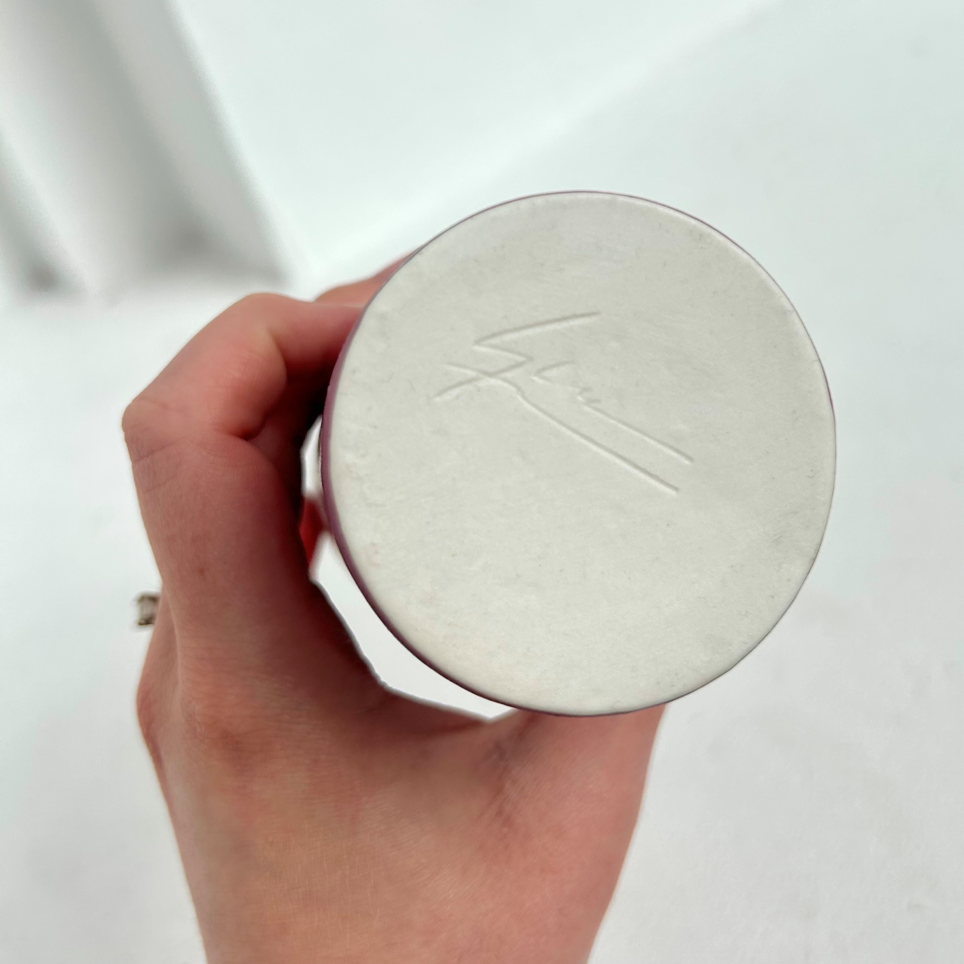 Hand holding a white cylindrical container with a brand logo on a light background